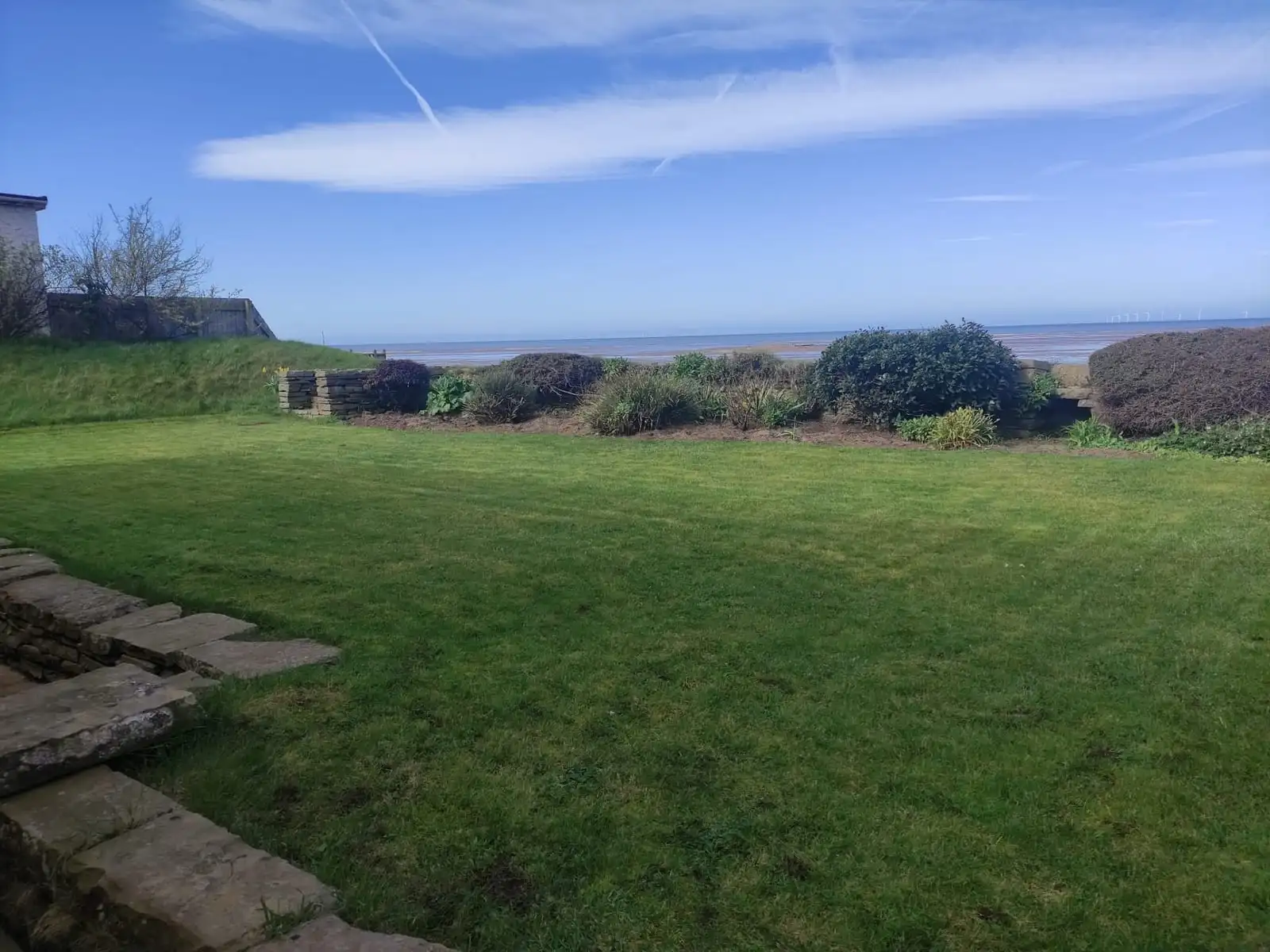 A coastal garden lawn looking out over the estuary, with a dry stone wall and wind turbines on the horizon