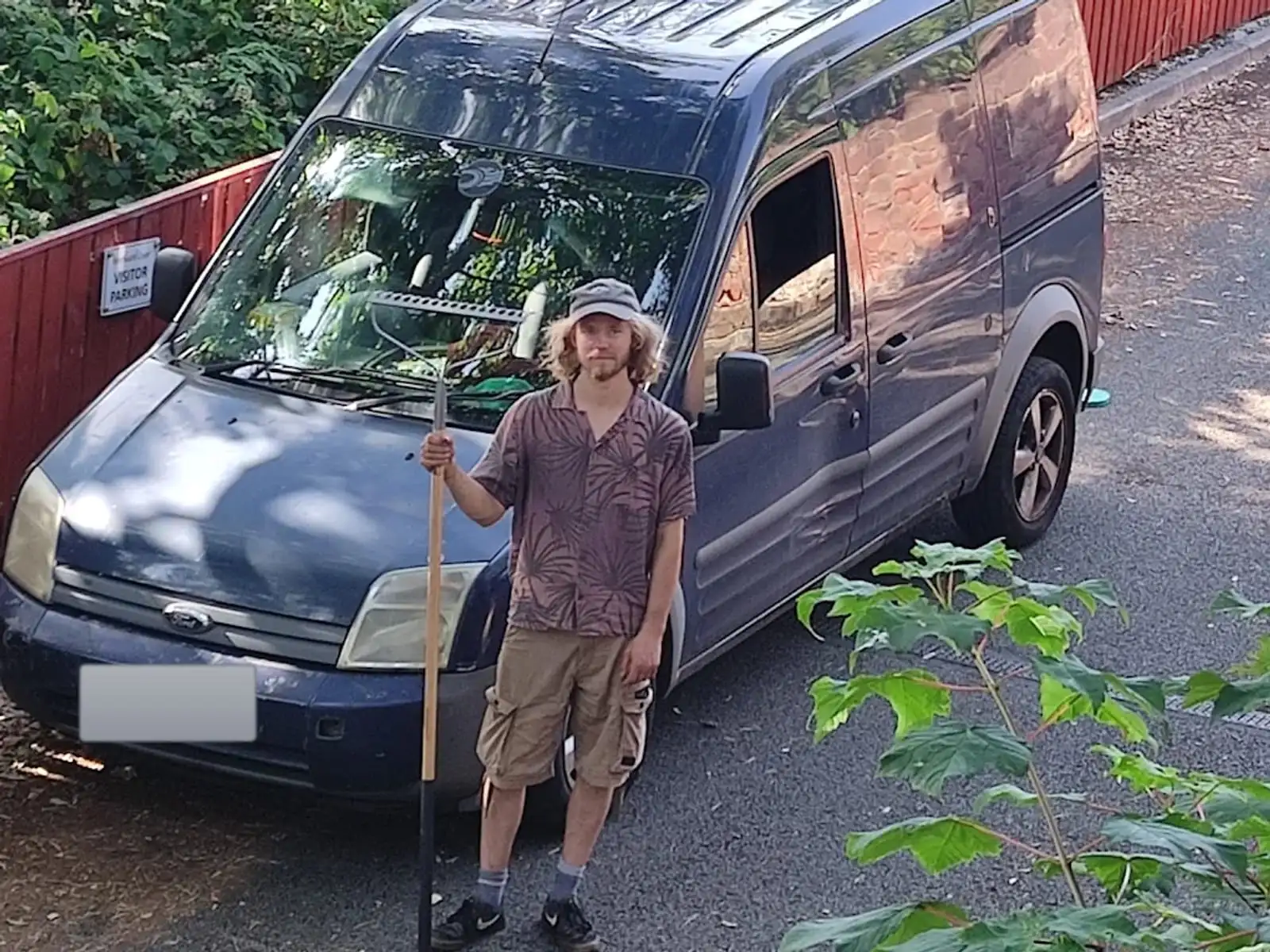 The gardener standing with a rake beside the Shangri-La Gardening van on a shady lane